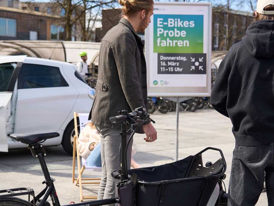 Vergrösserte Ansicht: Menschen mit Velo auf dem ETH Gelände und Plakat im Hindergrung zum Showcase Bikesharing