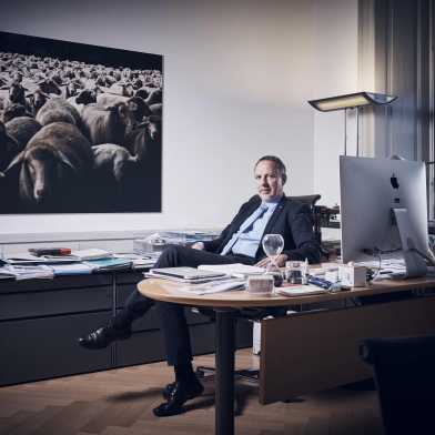Detlef Günther sitzt im Anzug in seinem Büro. Im Hintergrund ist ein Bild von einer Schafsherde zu sehen.
