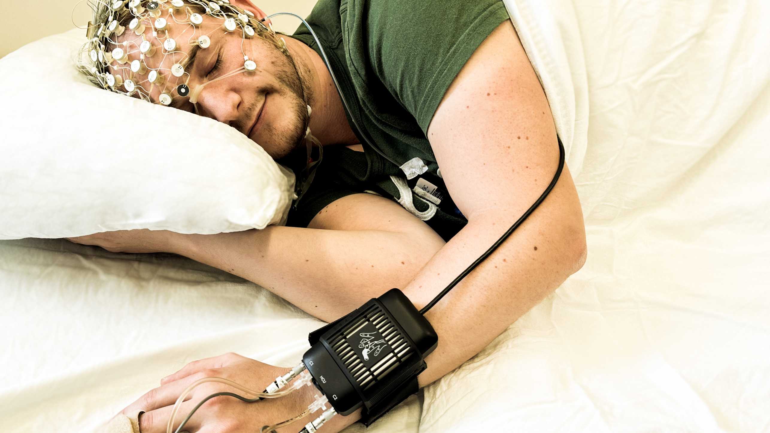 Mann liegt im Bett, auf dem Kopf sitzen Sensoren, am Handgelenk ein Kasten mit Drähten