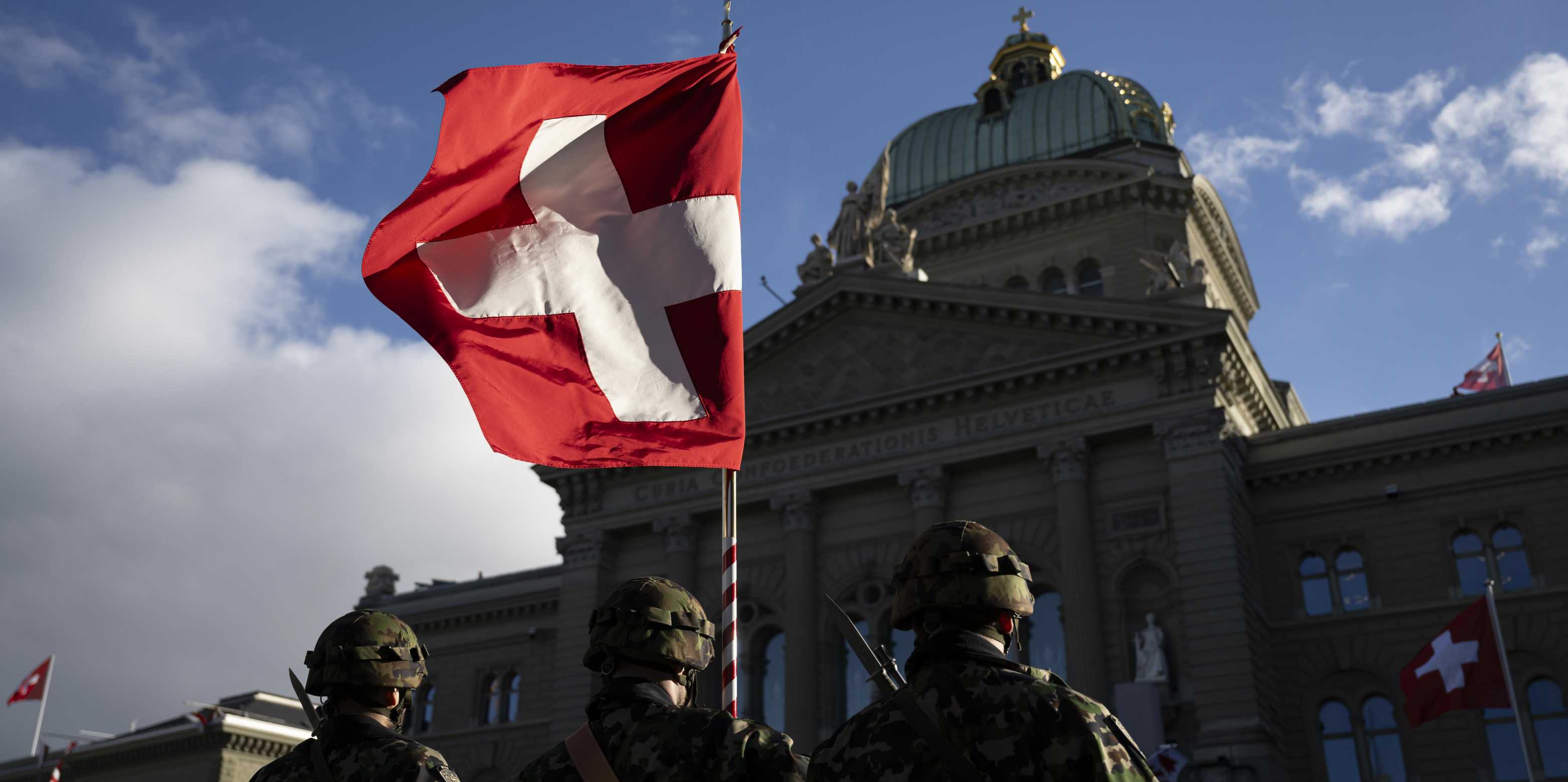 Drei Personen in Militärkleidung stehen vor dem Bundeshaus mit einer Fahne der Schweiz.