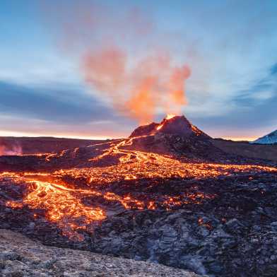 Un vulcano erutta sullo sfondo, la lava incandescente scorre sulla roccia scura in primo piano