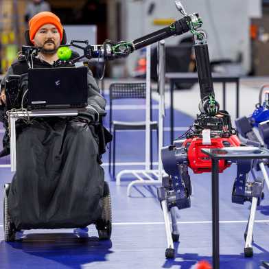 Samuel Kunz sitzt im Rollstuhl und ein Roboterhund führt ihm einen Apfel zum Mund.