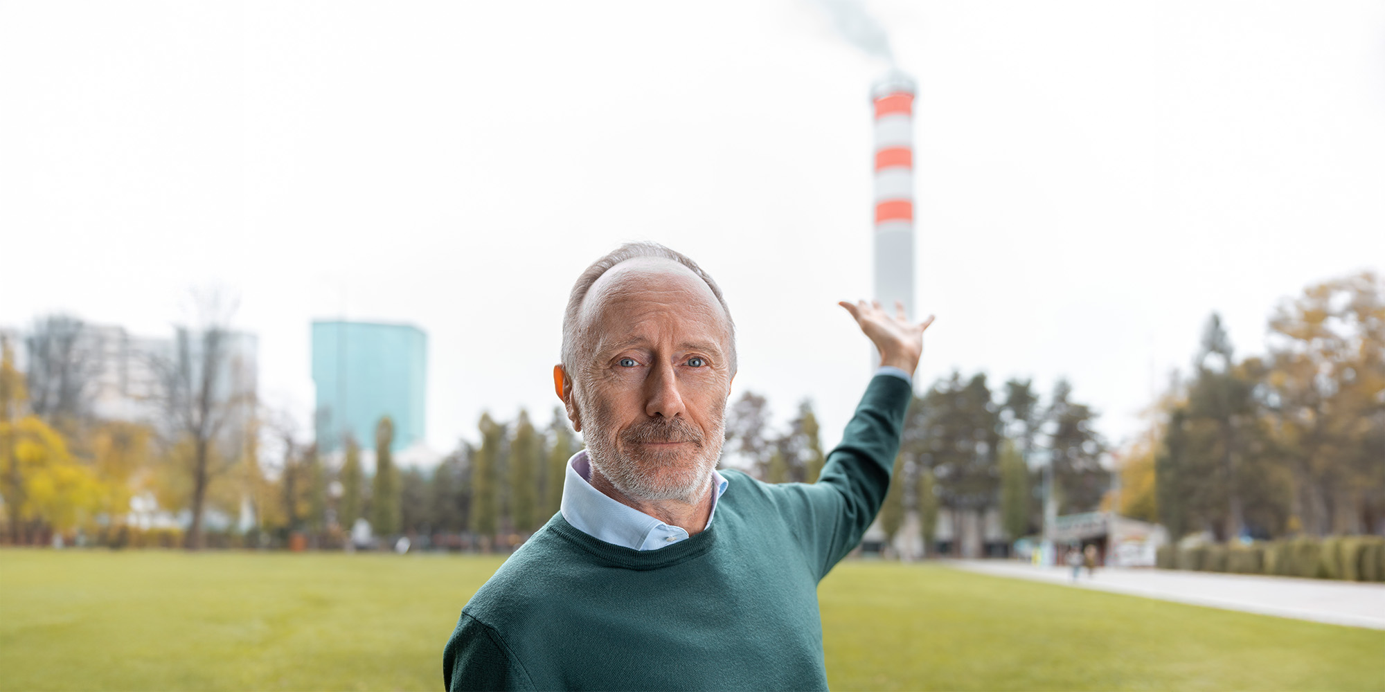 Marco Mazzotti schaut in die Kamera und deutet mit der einen Hand auf den Kamin eines Heizkraftwerks