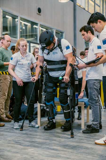 Vergrösserte Ansicht: Person geht im Exoskelett mit Krücken, umringt von Studierenden und Publikum
