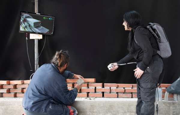 A bricklayer places a brick, a woman points a camera at it