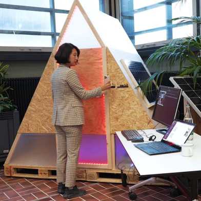 A woman stands in front of a triangular box with laptop and solar panel