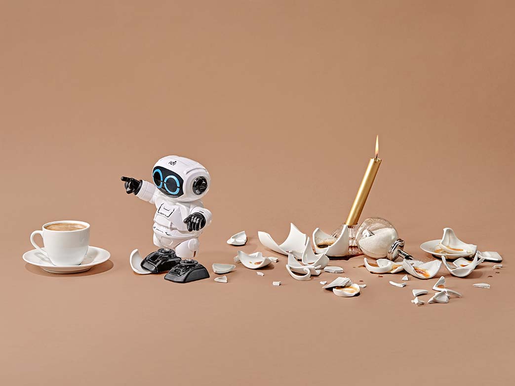 A mini robot next to a broken coffee pot, a coffee cup, a candle and a sugar dispenser
