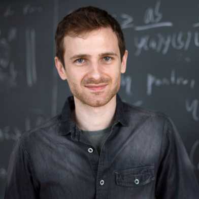 Oliver Janzer stands smiling in front of a written blackboard 