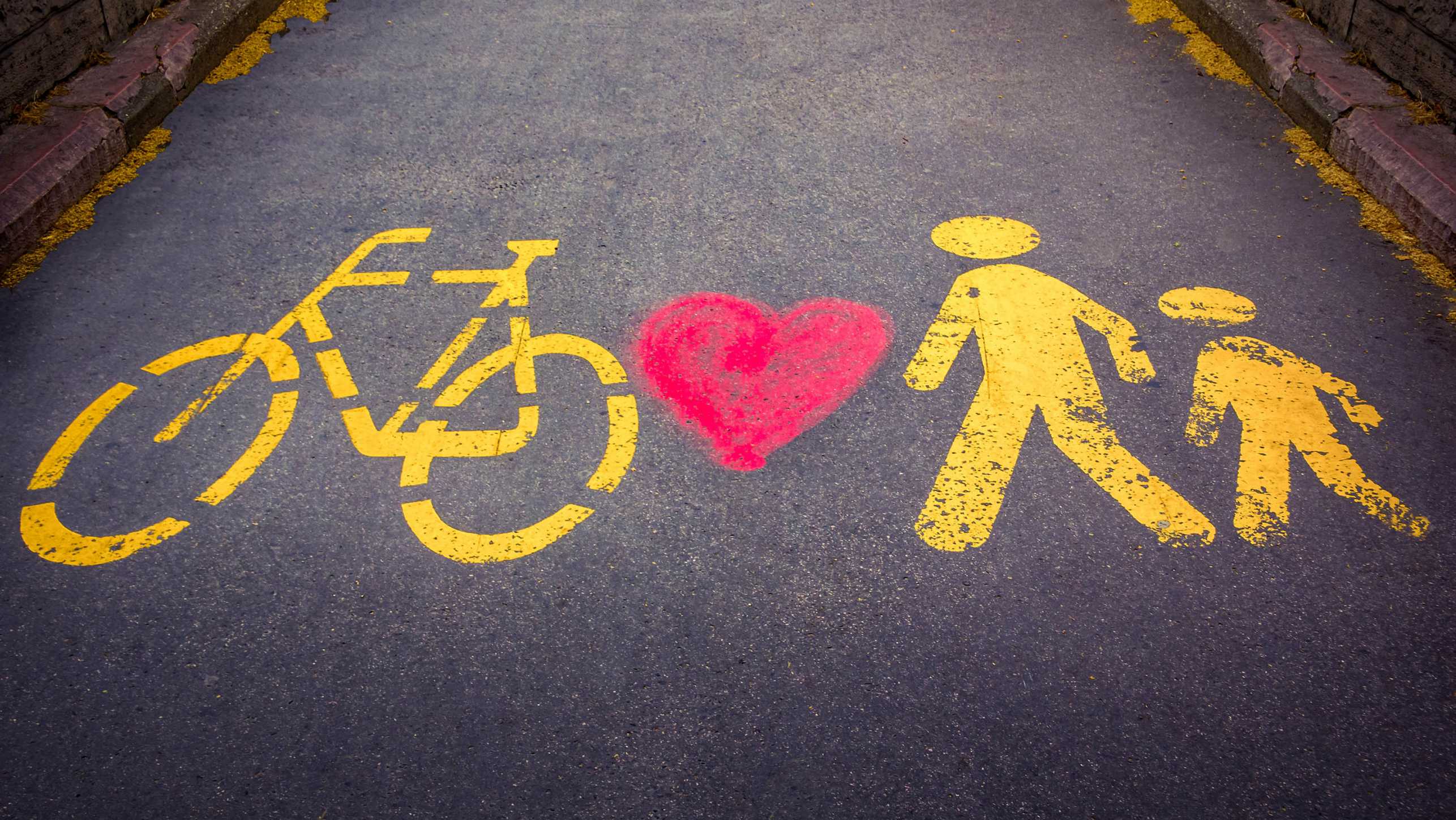On a paved path, the pictograms of a bicycle and two people are spray-painted in yellow with a red heart between the bicycle and the people.