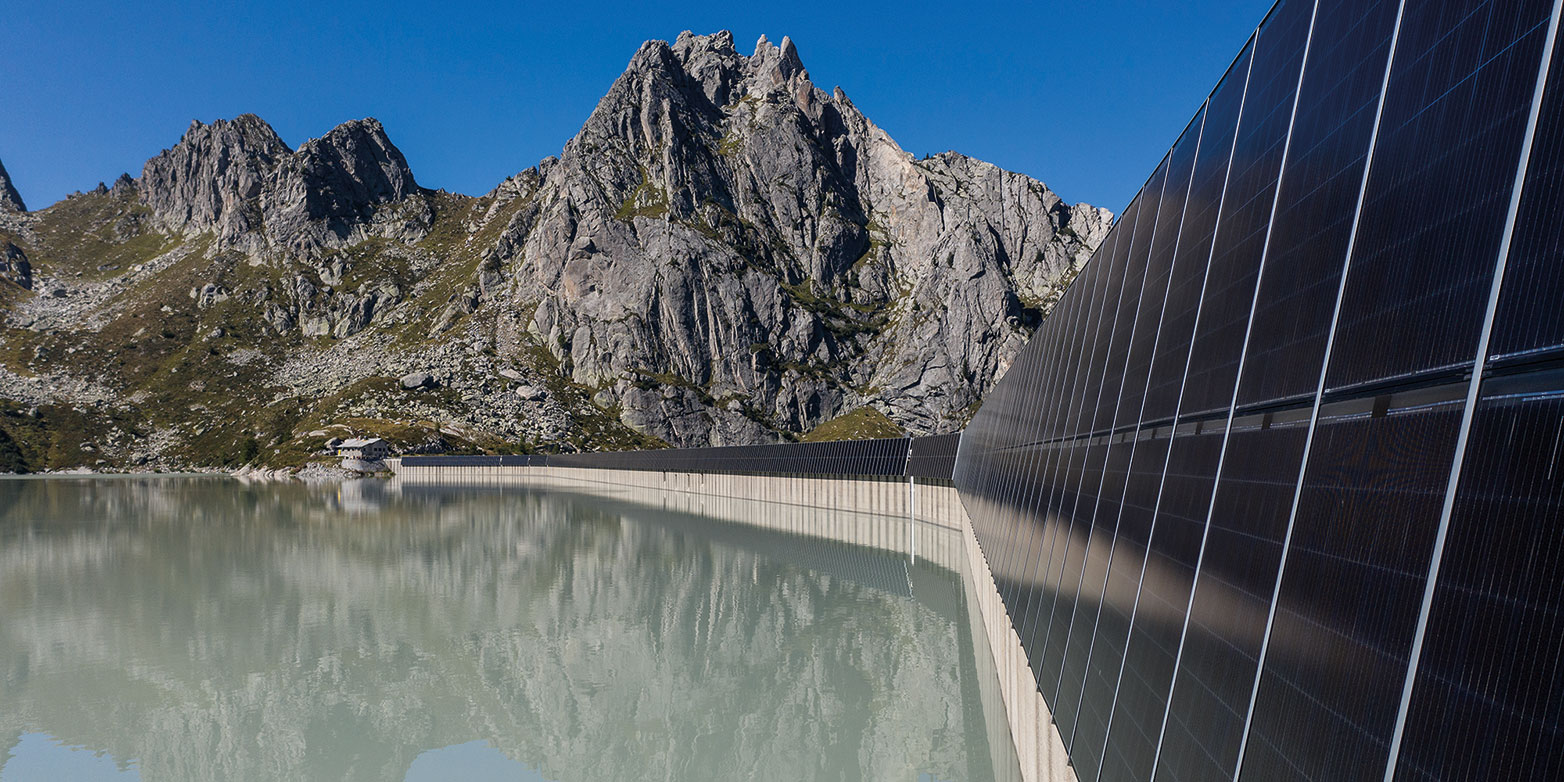 View over the Albigna dam filled with murky water, on whose walls solar panels are mounted. In the background, a craggy mountain rises into the blue sky.