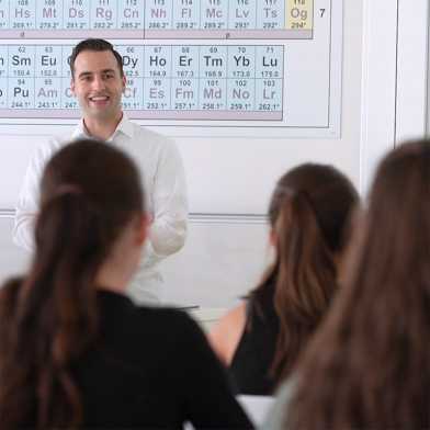 Lecturer is in front of a periodic table and teaches young women.
