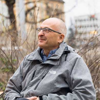 A man with a bald head, glasses and a grey The North Face jacket sits on a bench and looks into the distance.