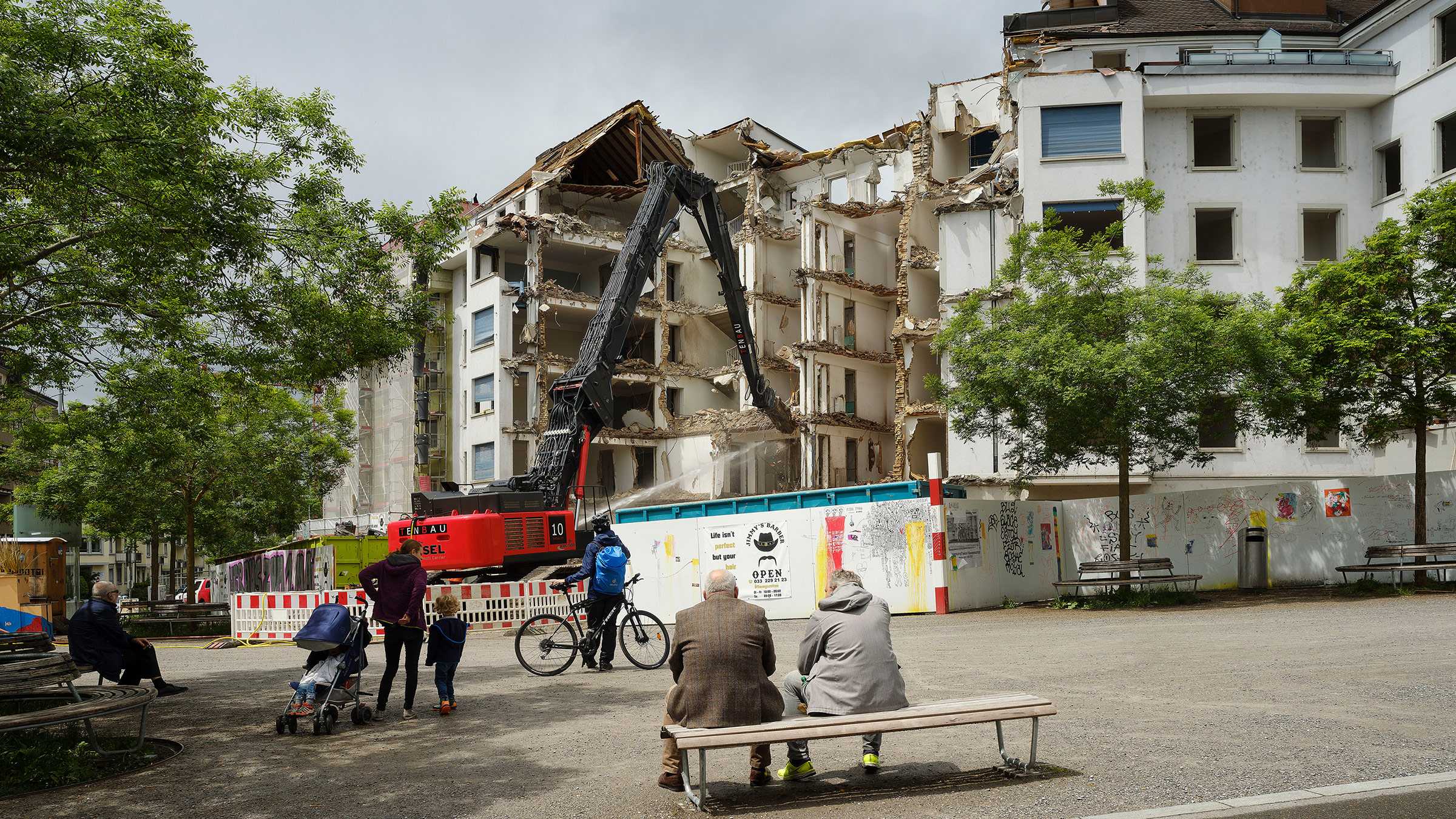 Two old people sit on a bench and watch as the building in front of them is demolished.