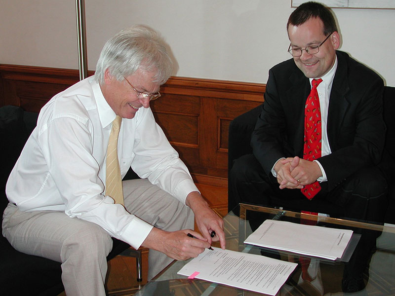 Enlarged view: Perich and Kübler at a table, both laughing, Kübler signing the financial regulations on a piece of paper.