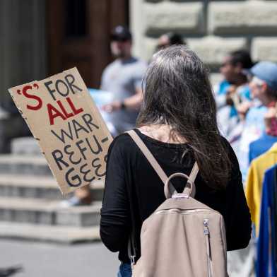 A woman holds a sign that says 'S' FOR ALL WAR REFUGEES.