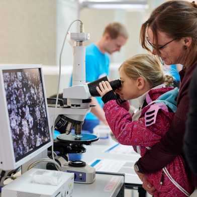 A little girl is held by her mother while she looks intently through a microscope.