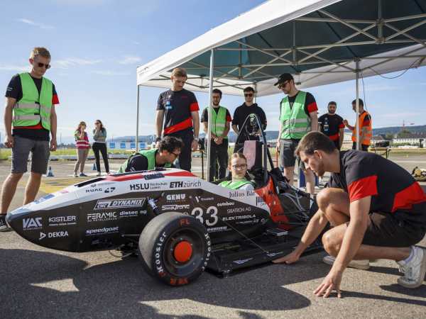 The pilot sits in the electric race car. The teammates stand around the vehicle and inspect it or carry out last preparations on it.