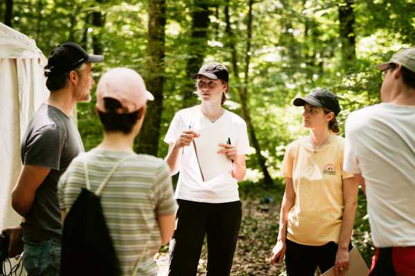 The five people stand in a circle in the forest and listen to the theoretical input.