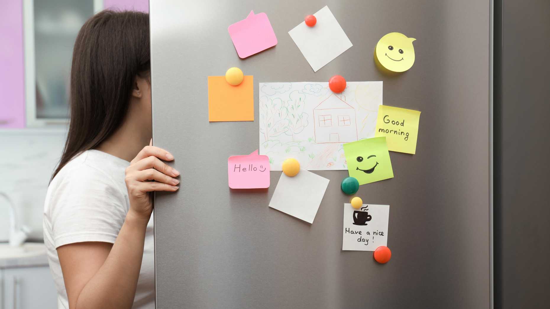 A woman opens the fridge and looks inside. The fridge door is covered in magnets and post-its