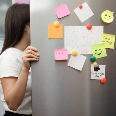 A woman opens the fridge and looks inside. The fridge door is covered in magnets and post-its