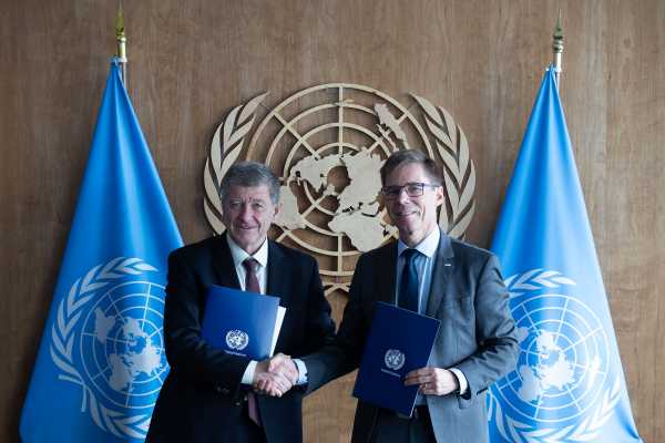 Guy Ryder, pictured left, and Joël Mesot shaking hands. They are each holding a blue folder, with the UN emblem in the background