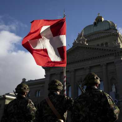 Three soldiers in military uniform stand in front of the Federal Palace with a Swiss flag.