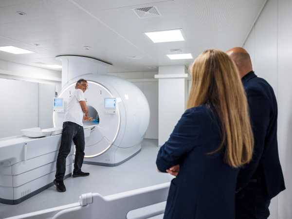 A man stands next to an MRI machine and sexplains the machine to a man and a woman who listen
