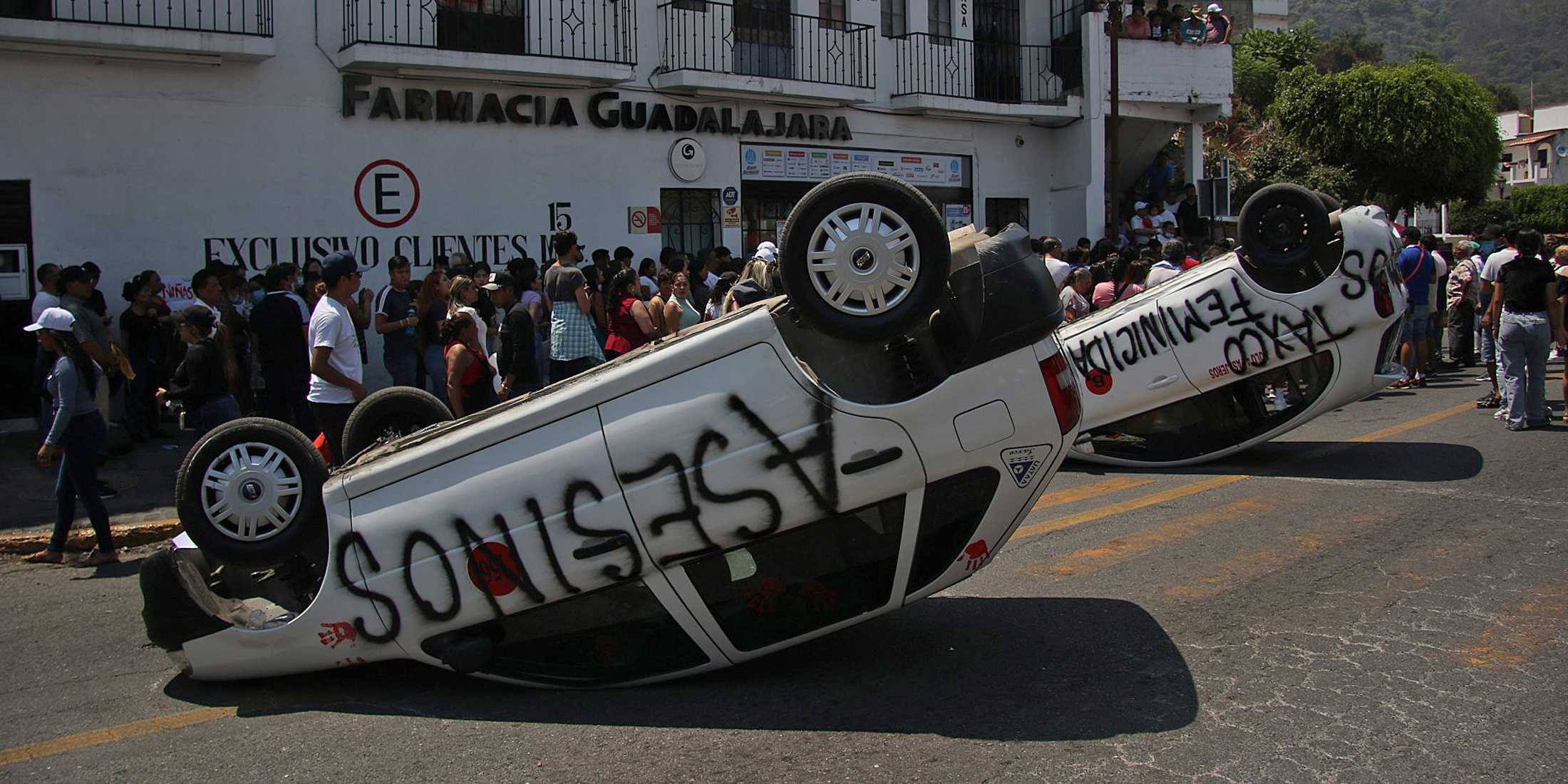 Two cars lie upside down on the road and are spray-painted with words like "Asesinos". Civilians are recognizable in the background.