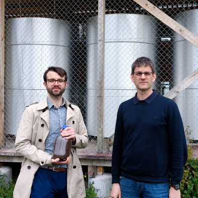 ETH researchers Samuel Heiniger (left, with a glass of iron ore) and Professor Wendelin Stark in front of the three iron reactors on the Hönggerberg campus of ETH Zurich. 