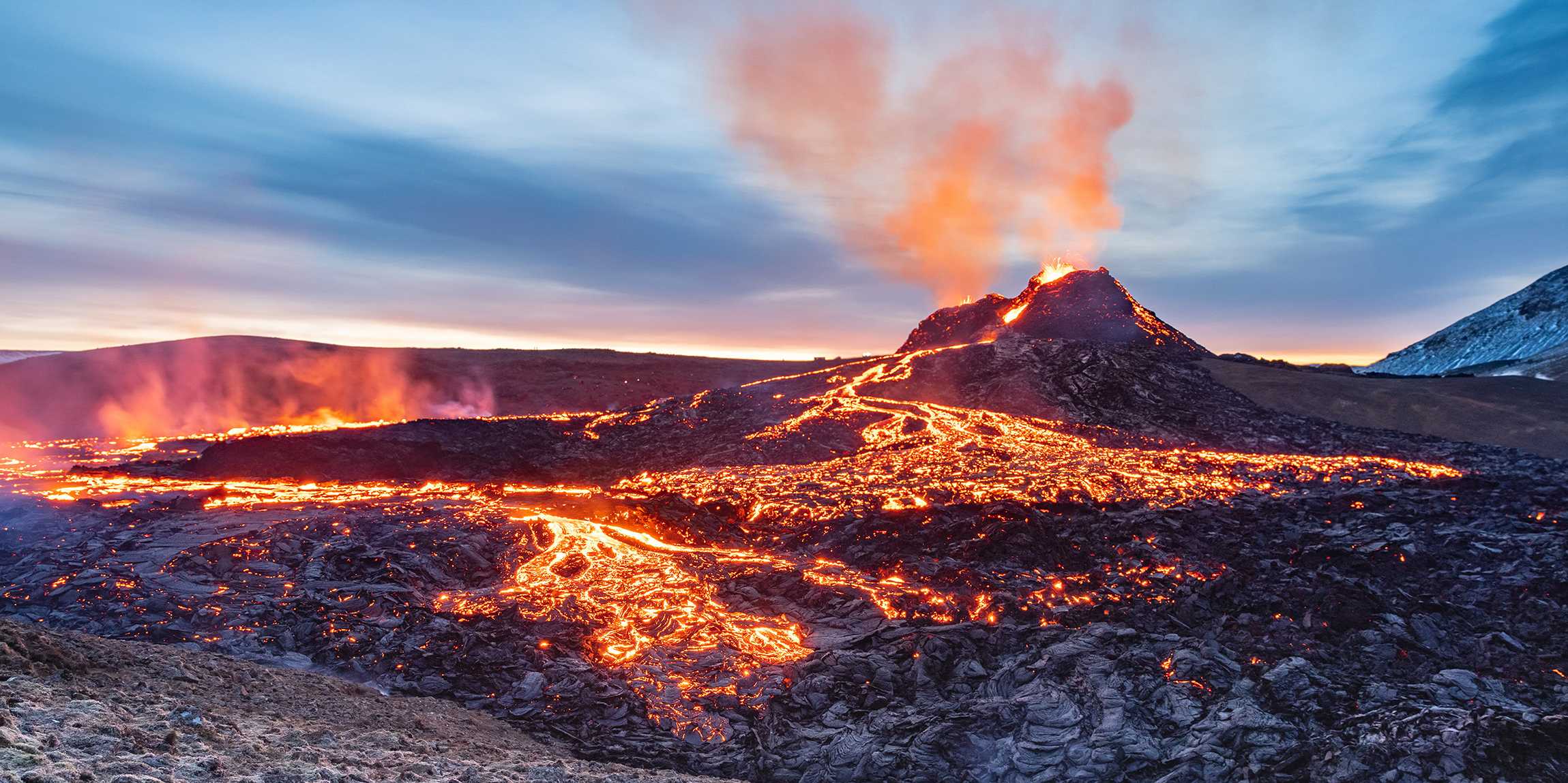 A volcano erupts in the background, glowing lava flows over dark rock in the foreground