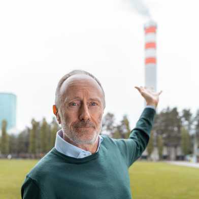 Marco Mazzotti looks into the camera and points with one hand to the chimney of a combined heat and power plant