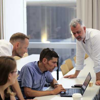 A man and a woman sitting by a table and two men standing. They are all looking at a laptop and discussing something.