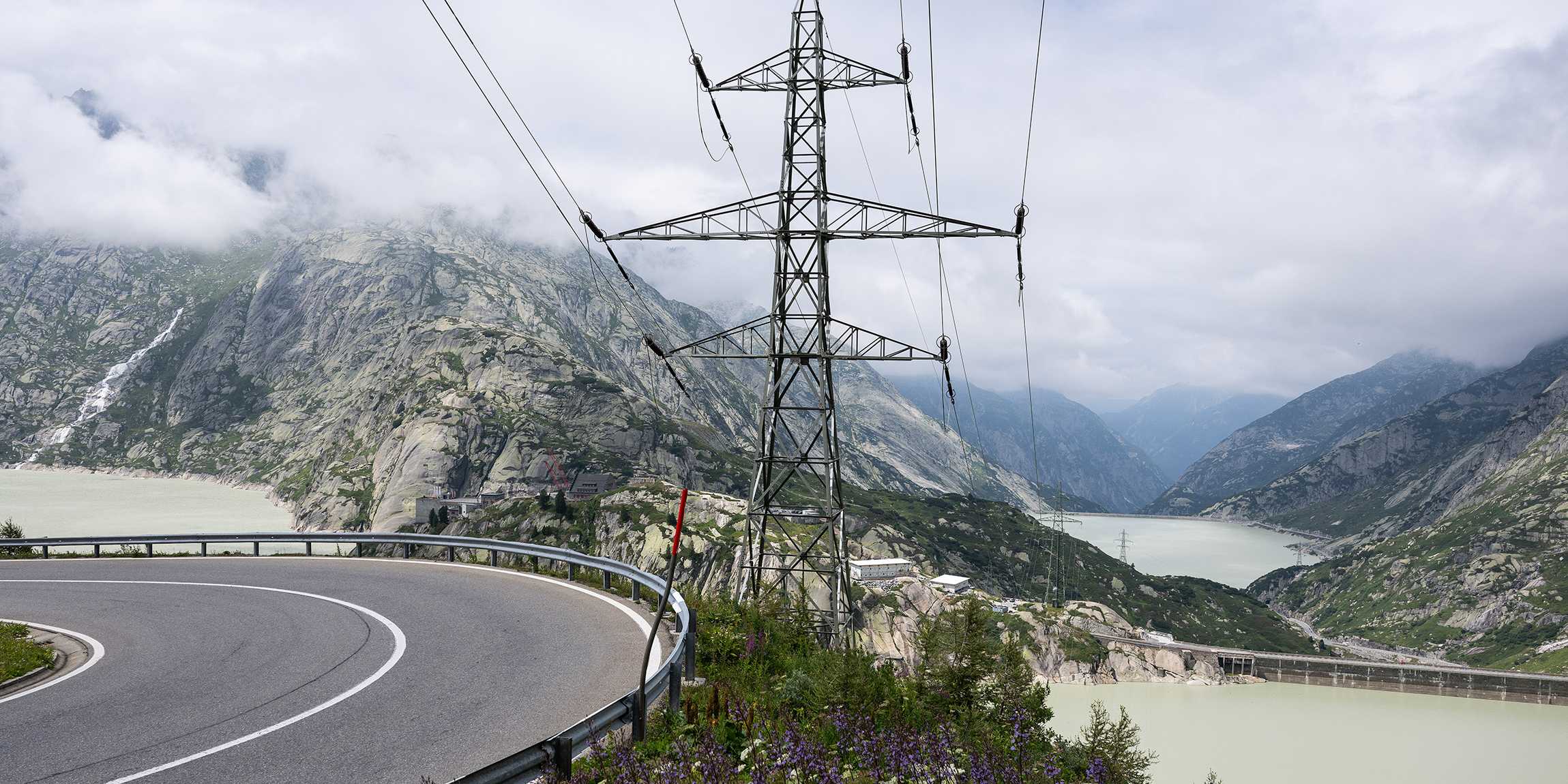 A road that leads past a water reservoir in the mountains. There is an electricity pylon right next to the reservoir.