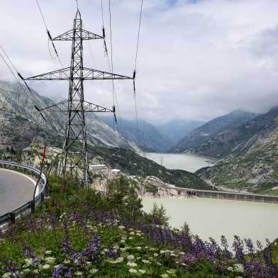 A road that passes a reservoir in the mountains. A power pylon with high-voltage lines is visible in the foreground.