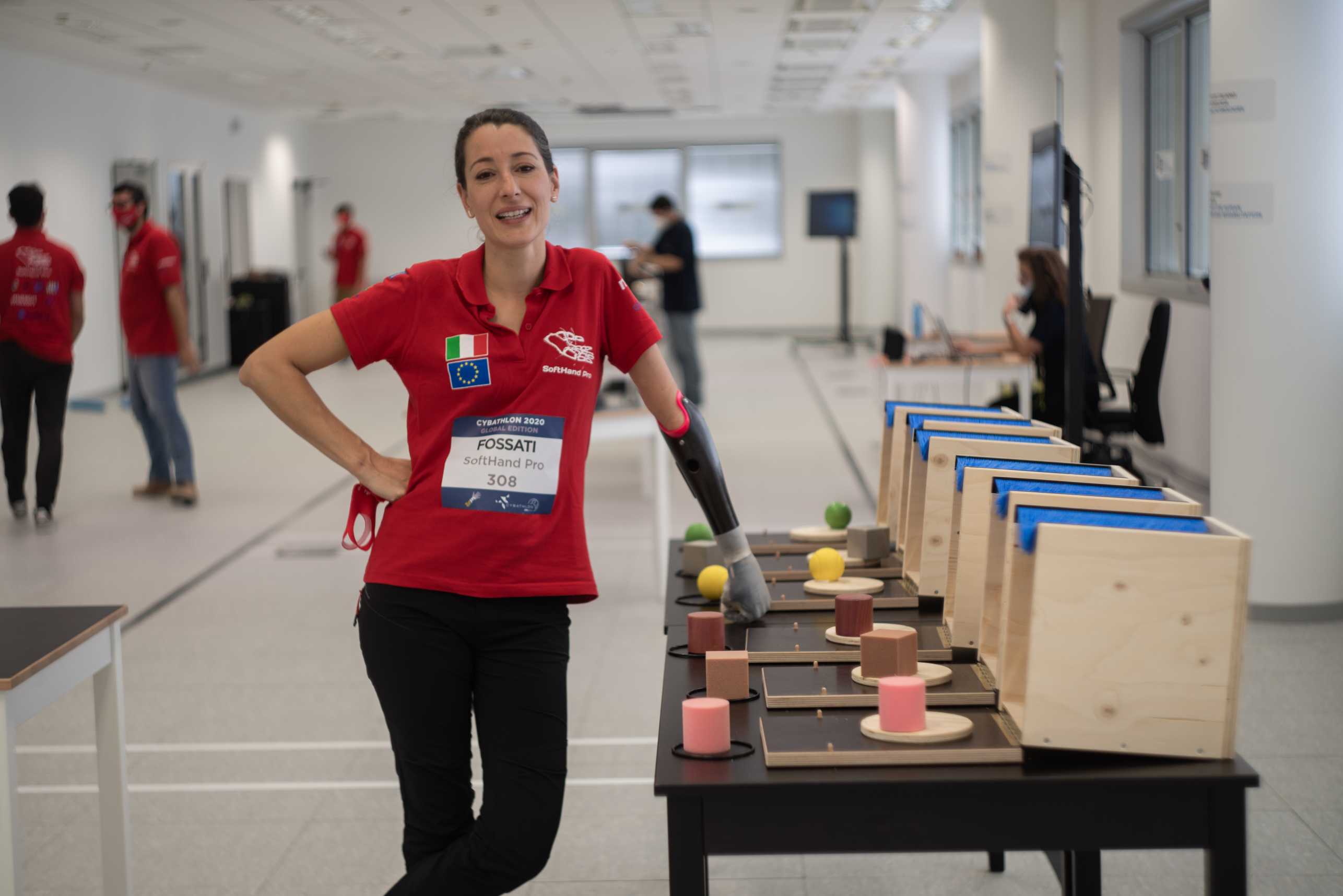 Maria Fossati, designer and researcher, standing next to what appears to be a table with CYBATHLON competition props. She is resting her prosthetic arm on the table.