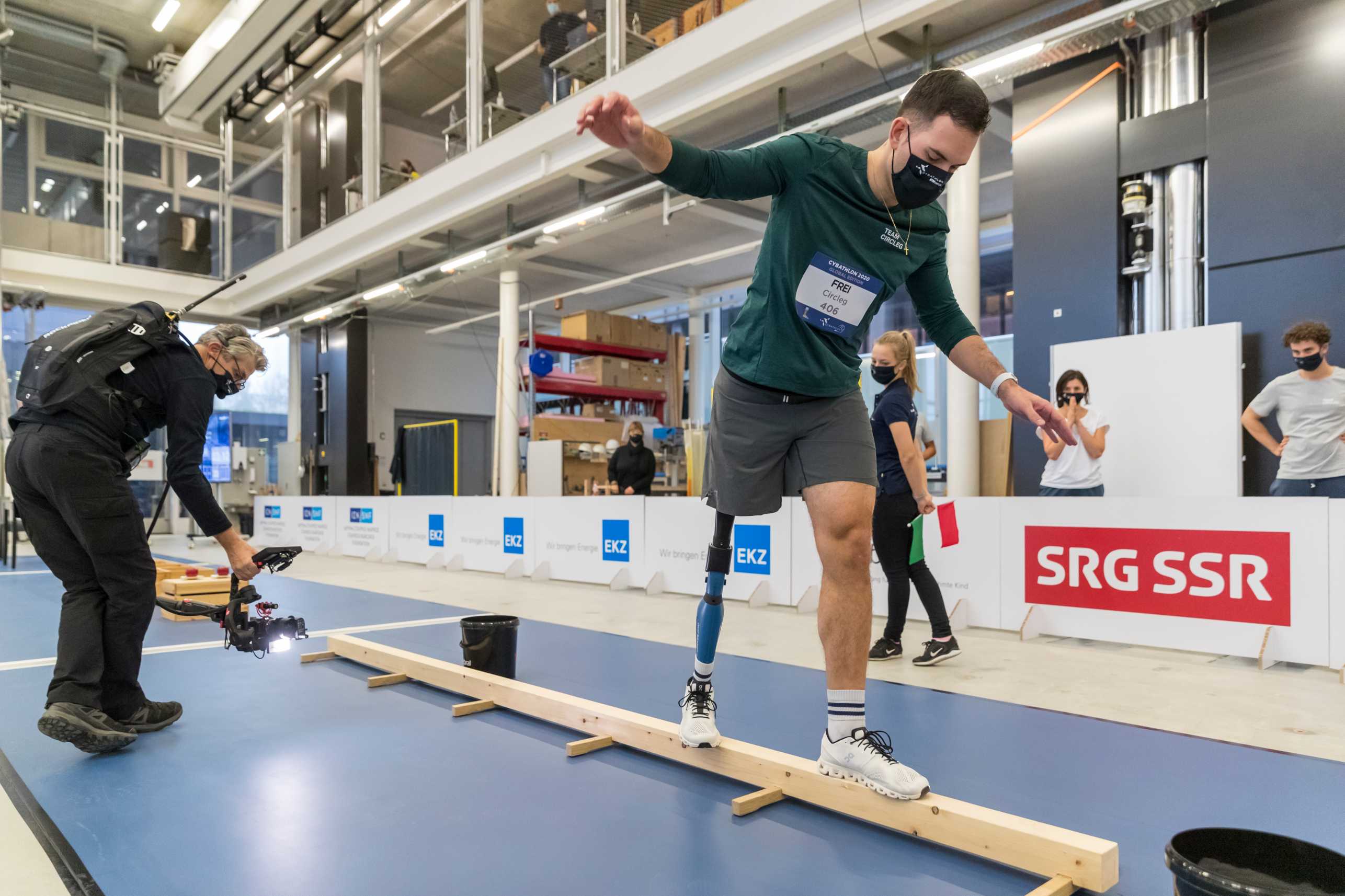 A man with a leg prosthesis on his right leg is balancing over a wooden obstacle. The obstacle is about two meters long and ten centimeters high. A camera man is behind him filming.