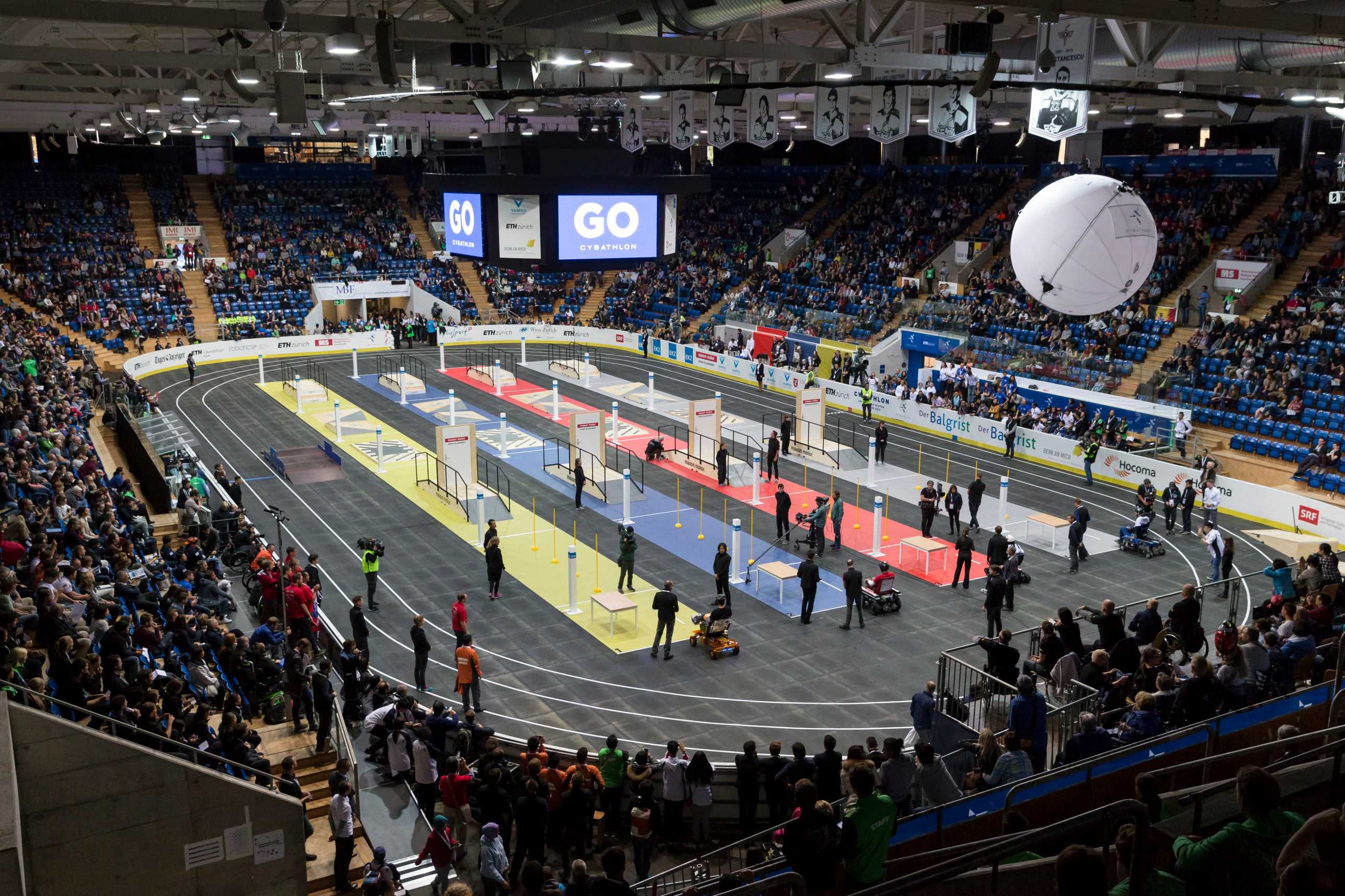 A stadium filled with people. In the middle of the stadium, CYBATHLON races are in place. The crowd is cheering the athletes on.