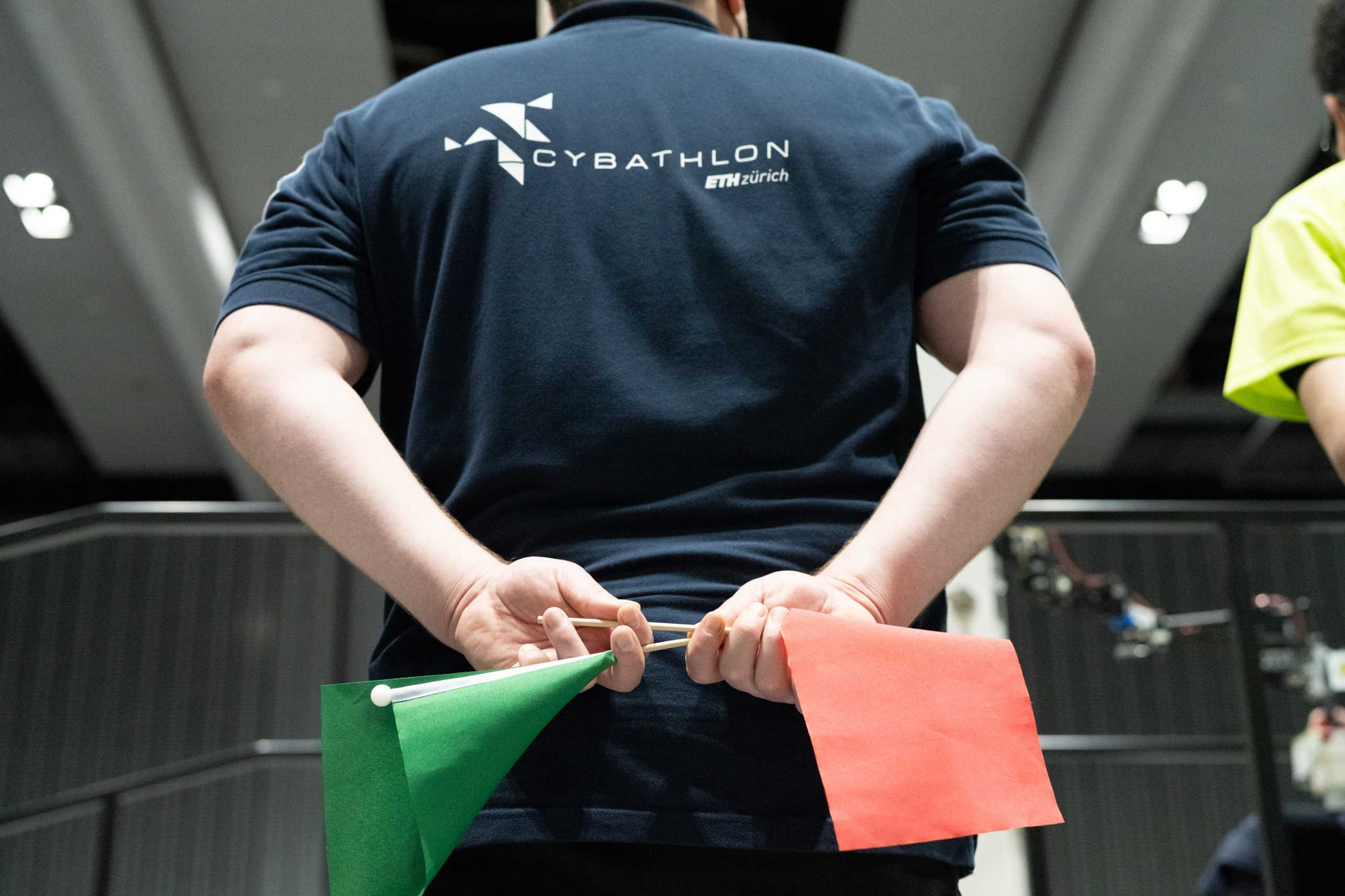 A man is seen from behind. He is wearing a Tshirt with the CYBATHLON and ETH Zurich logo. He is holding two flags; a green one and a red one. He appears to to a referee for the CYBATHLON games.