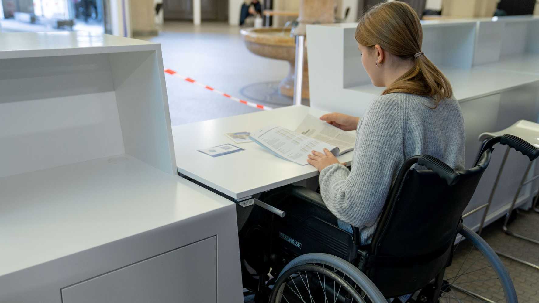 Vergrösserte Ansicht: Eine junge Frau sitzt in einem Rollstuhl an einem barrierefreien Registrationsdesk.