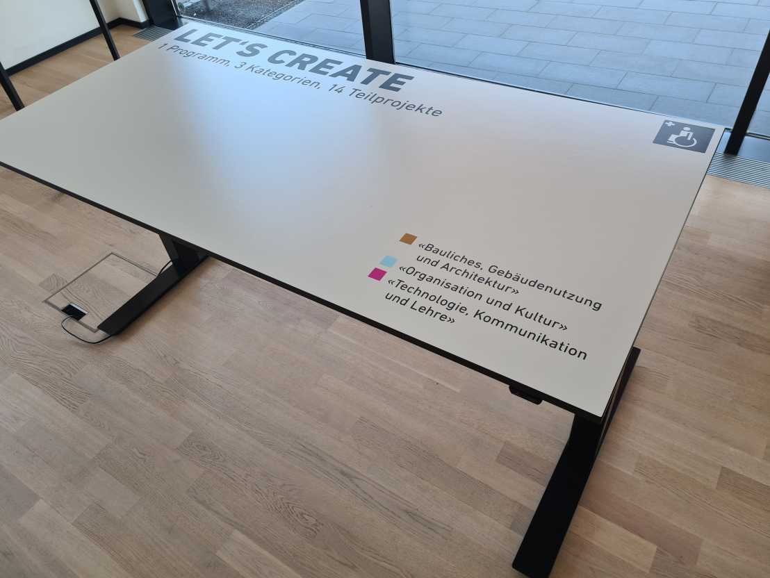 The surface of the electrically adjustable desk features the words “Let’s create. 1 programme, 3 categories, 14 sub-projects”. An explanation: the programme is called “Barrier-Free at ETH Zurich”. The three categories are “Construction, building use and architecture”, “Organisation and culture” and “Technology, communication and teaching”.