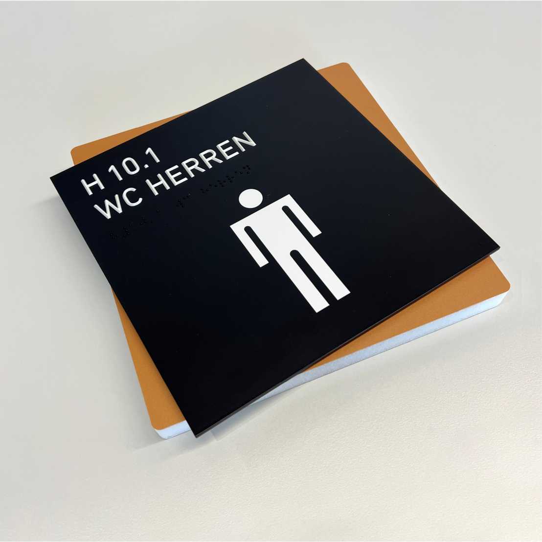 square door sign for a men’s bathroom with a white figure on a black background as well as raised lettering and Braille