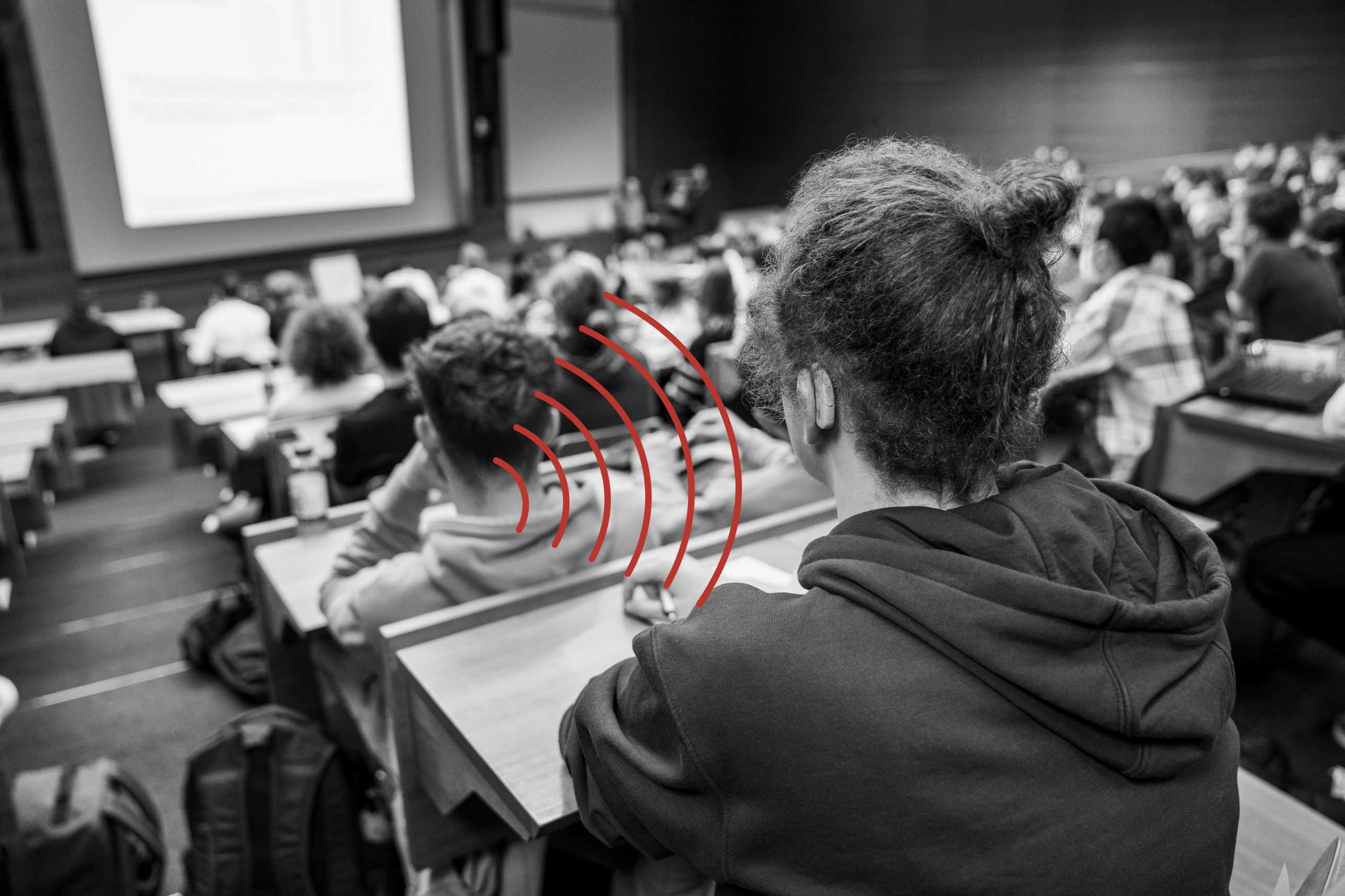Enlarged view: A student with a hearing aid sits in a lecture hall full of students. The hearing aid picks up the acoustic signals via an induction loop.