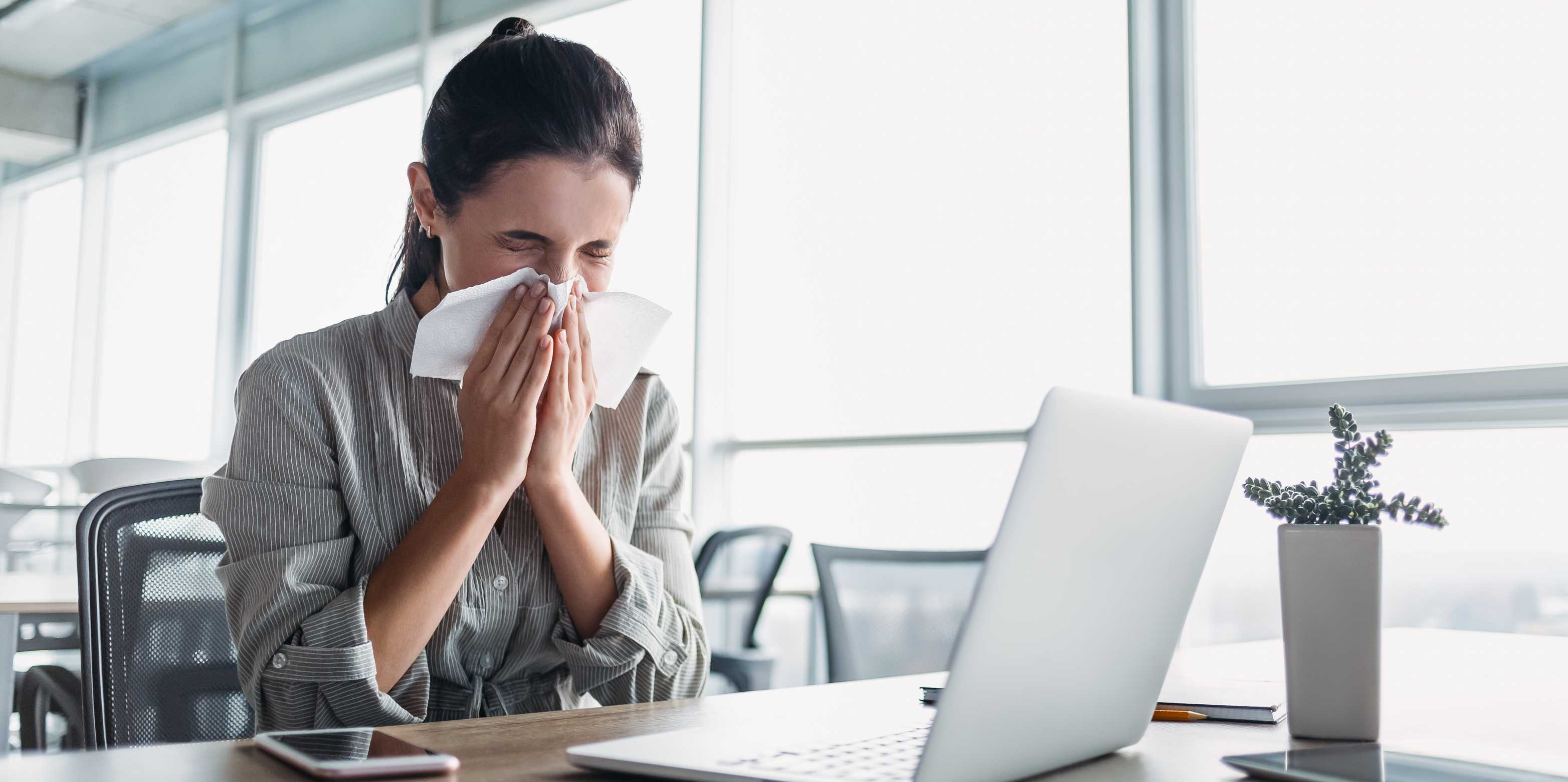 A woman sits in front of a laptop and blows her nose.