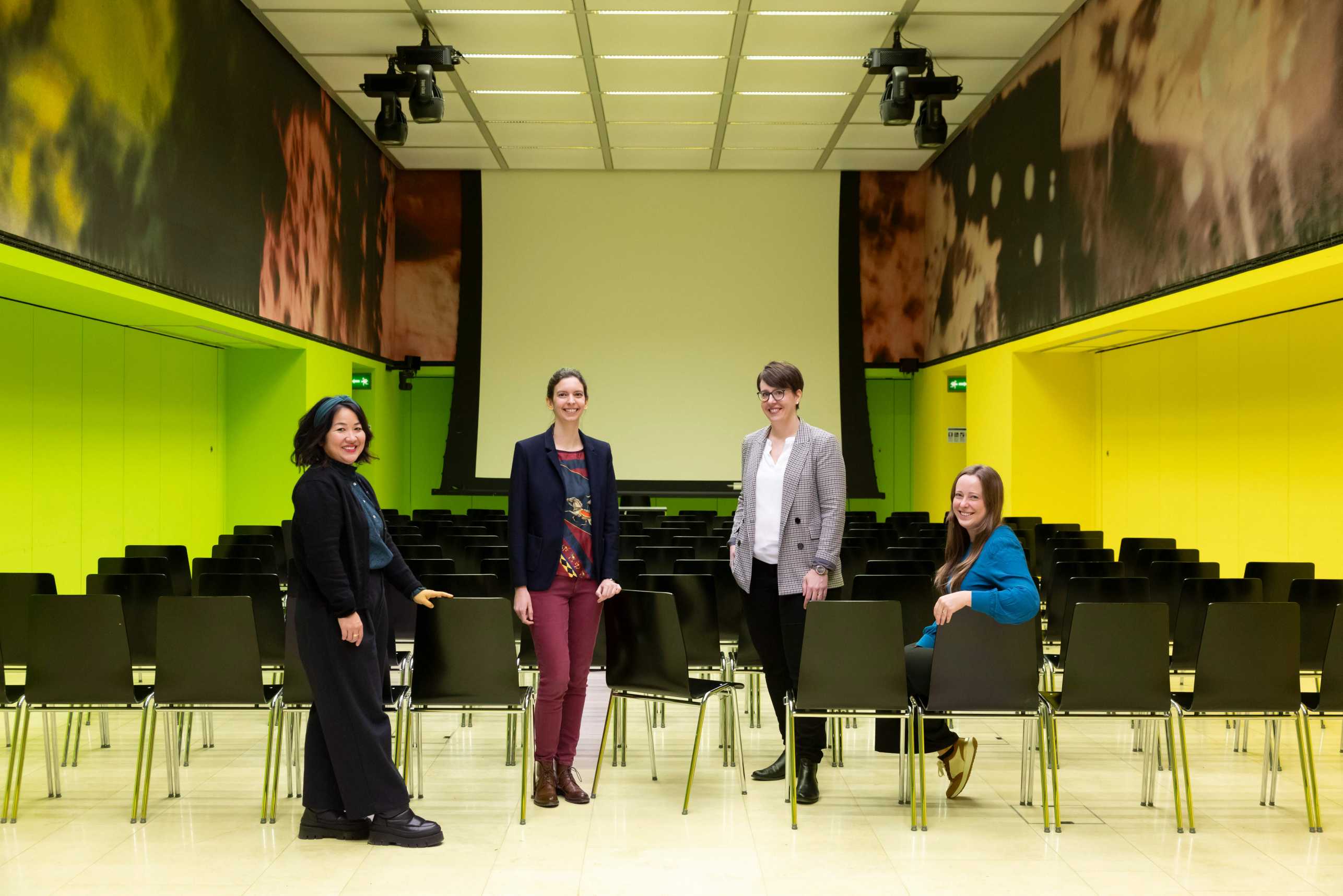 Four people are in a room with green walls and lots of black chairs