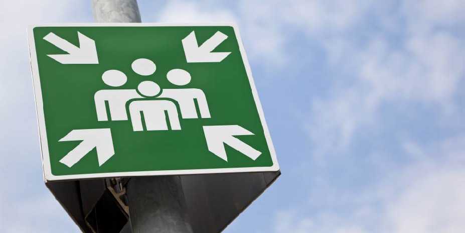 In the photo, you can see a large green sign with a pictogram representing a person. The sign forms a square and arrows point to the person from all four corners. It is attached to a column and the background is a blue sky with clouds.