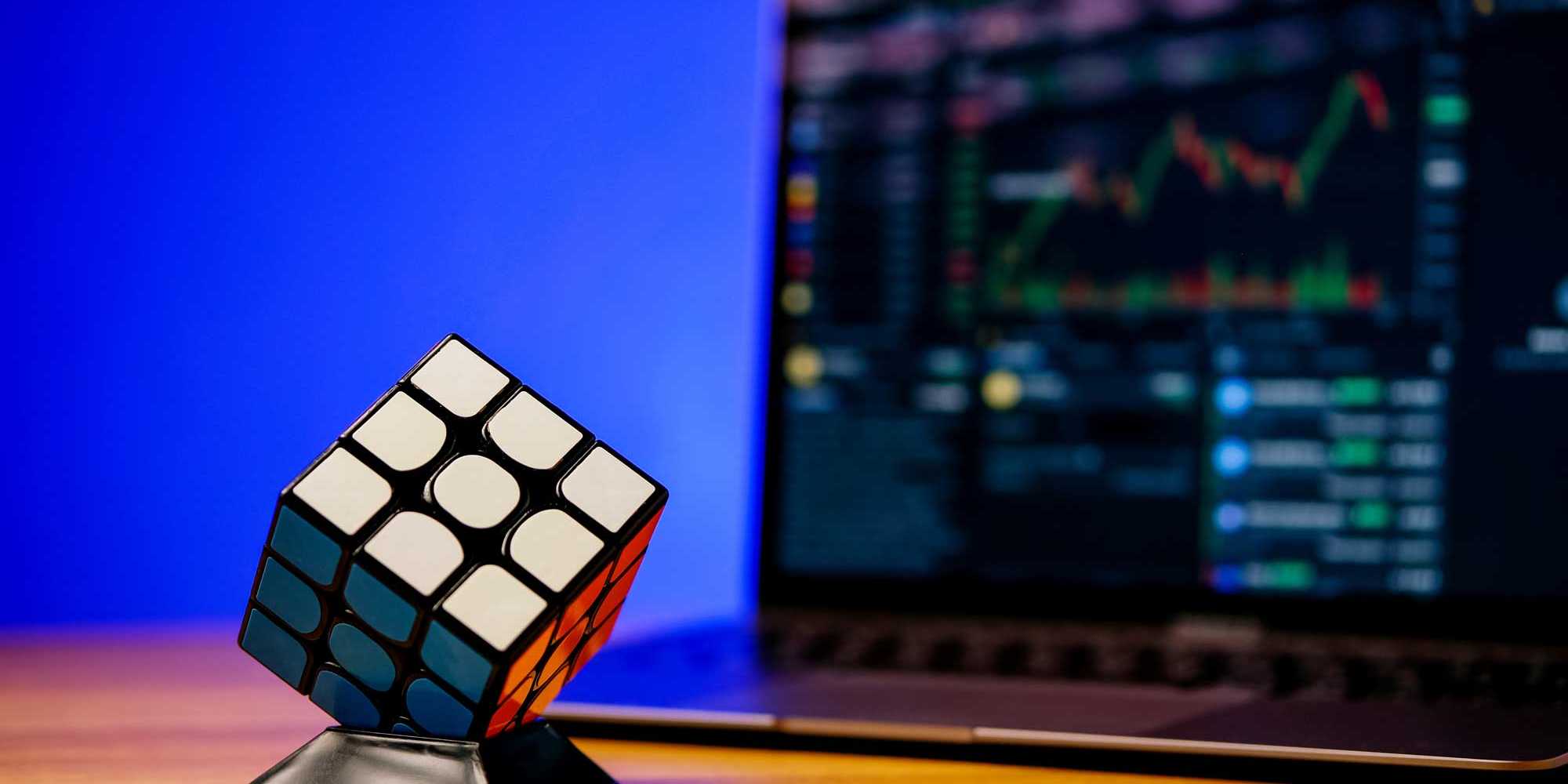 Photo shows a Rubik's cube in front of a screen with financial data and curves.